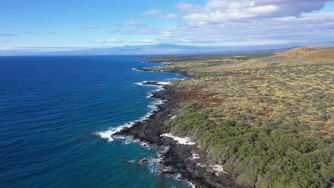 Aerial-view-of-the-Hawaiian-coastline-with-rocky-shores,-blue-ocean,-and-rolling-hills-under-a-clear-sky