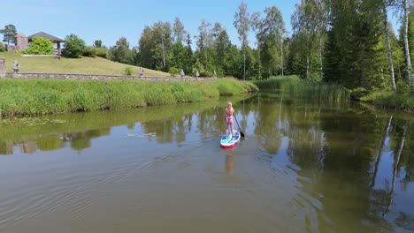 Volando-Por-Encima-De-Una-Joven-De-Pie-Remando-En-Una-Tabla-De-SUP-En-Un-Estanque-En-Verano