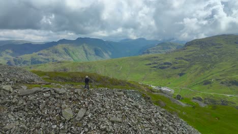 Lone-mountain-walker-stood-on-crumbling-mountainside-looking-towards-distant-mountains-under-overcast-sky