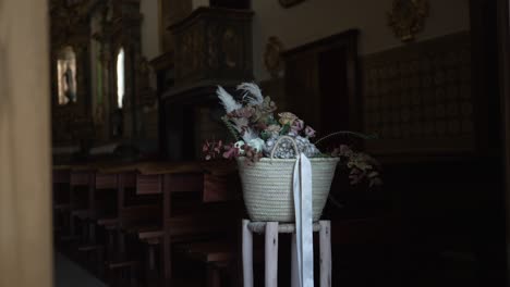 Cesta-Rústica-Con-Flores-Y-Vegetación-En-Un-Stand-Dentro-De-Una-Iglesia-Con-Poca-Luz.