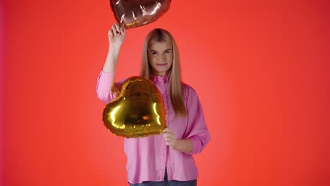 Hübsche-Blonde-Frau-Schwenkt-Goldene-Herzballons-Und-Wirft-Sie-Dann,-Farbenfrohe-Studioaufnahme