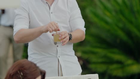 Man-in-a-white-shirt-stirring-a-drink-at-an-outdoor-event-with-greenery-in-the-background