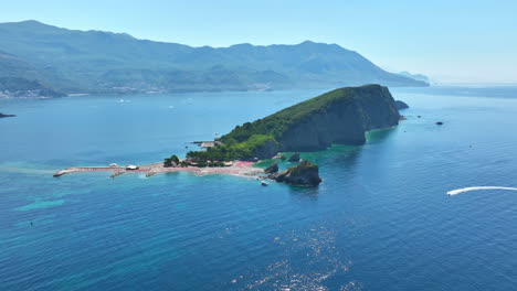 Drone-rotating-away-from-the-Sveti-Nikola-Island,-sunny,-summer-day-in-Montenegro