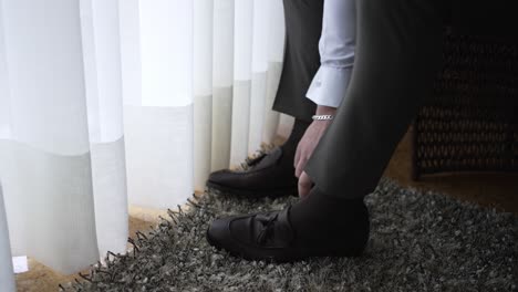 Hombre-Ajustando-Sus-Zapatos-Formales-Negros-Mientras-Está-Sentado-En-Una-Silla-Cerca-De-Una-Ventana