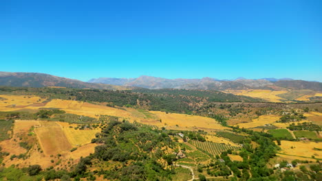 Ausgedehnte-Luftaufnahme-Von-Sanften-Hügeln-Und-Landwirtschaftlichen-Feldern-Mit-Einer-Bergkette-Im-Hintergrund-Unter-Einem-Klaren-Blauen-Himmel