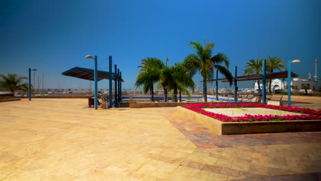 Wide-angle-view-of-a-seaside-promenade-with-palm-trees,-flowerbeds,-and-modern-amenities,-overlooking-the-marina-under-a-bright-blue-sky