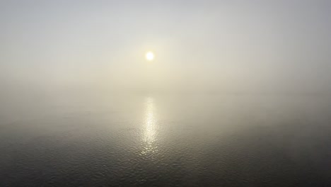 Una-Amplia-Toma-Estática-Del-Amanecer-En-Medio-De-La-Niebla-Matutina-Tomada-Sobre-Un-Cuerpo-De-Agua-Que-Fluye-Con-El-Reflejo-Del-Sol