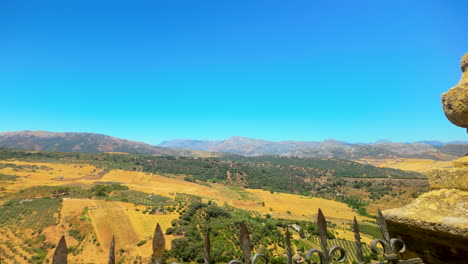 Scenic-view-of-a-vast-valley-with-fields-and-mountains-in-the-distance,-seen-from-a-stone-balcony-with-wrought-iron-railing