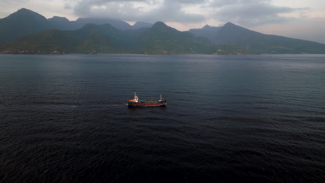 Seafaring-ship-navigating-open-sea-off-Bali-mountainous-coastline,-aerial-view