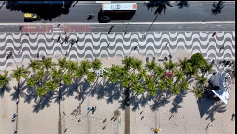 Drone-footage-captures-Ipanema-Beach-from-above,-showcasing-people-playing-sports,-strolling-along-the-sidewalk,-and-vehicles-passing-by,-all-framed-by-the-iconic-palm-trees-and-boardwalk