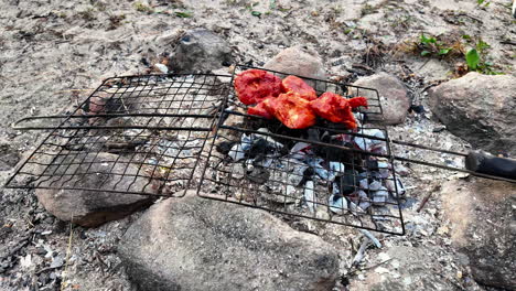 Someone-is-laying-meat-on-a-grill-to-bbq-it-on-the-beach-in-the-sand,-slow-motion-closeup