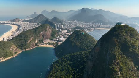 Breathtaking-aerial-footage-of-Rio-de-Janeiro’s-Sugarloaf-Mountain-with-a-cable-car-ascending,-set-against-the-cityscape-and-pristine-beaches,-captured-in-stunning-clarity-during-midday