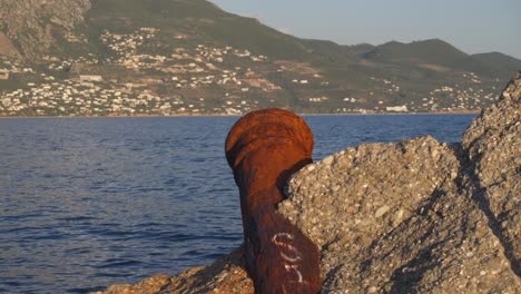 Toma-Estática-Media-De-Un-Viejo-Cañón-Vintage-Oxidado-Plantado-En-Un-Muelle-Rocoso-En-Kalamata,-Con-La-Montaña-Taigetos-Al-Fondo-4k