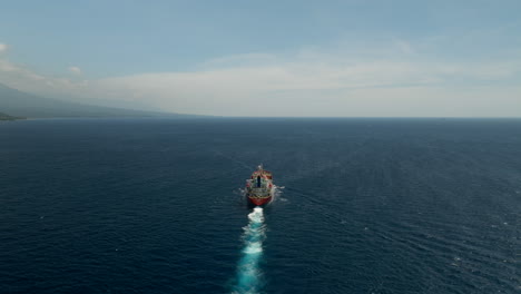 Red-tanker-ship-ploughing-through-blue-ocean-near-coastline,-rear-aerial-arc