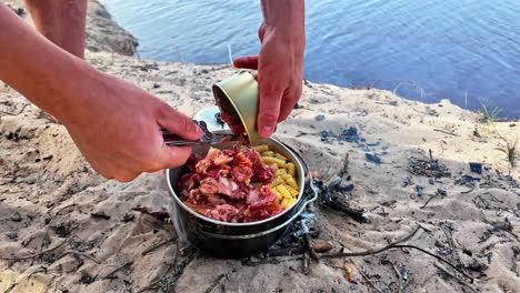 Male-hands-put-bacon-in-a-pot-with-noodles-on-the-cooking-fire-on-the-beach-in-the-sand,-slow-motion-close-up