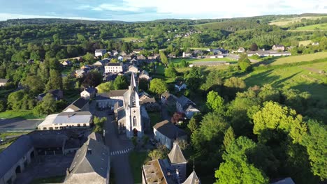 A-picturesque-aerial-view-of-the-Belgian-village-of-Ferrières,-showcasing-a-serene-rural-landscape-with-a-prominent-church-steeple,-traditional-houses,-surrounding-rolling-hills,-and-a-farm