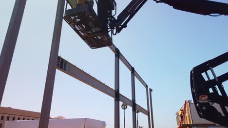 Gimbal-booming-down-shot-of-a-construction-worker-in-a-raised-bucket-lift-crane-at-a-building-site-in-West-Los-Angeles,-California