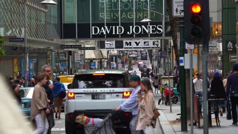 Shopping-precinct-featuring-David-Jones-and-Myer-flagship-stores-in-bustling-downtown-Melbourne-city,-with-pedestrians-crossing-and-busy-vehicle-traffic,-street-scene-of-Little-Bourke-Street