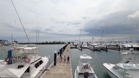 People-walk-back-towards-the-dock-on-the-pier-between-the-moored-hobby-and-sportfishing-ships-at-Marina-Pez-Vela-in-Quepos,-Costa-Rica