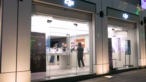 Pedestrians-stroll-past-DJI's-flagship-store,-known-for-its-drones,-video,-and-camera-gear,-on-a-Hong-Kong-night