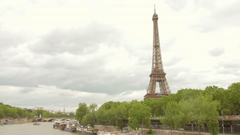 The-Eiffel-Tower-stands-tall-against-a-cloudy-sky-in-Paris,-surrounded-by-lush-green-trees