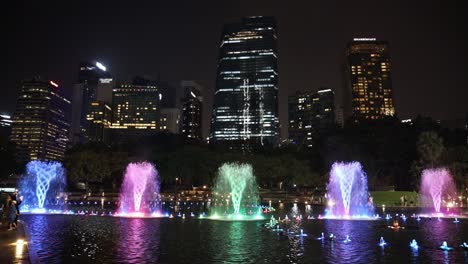 Five-water-spouts-in-various-colors-spraying-in-a-pond-in-front-of-KLCC-Petronas-Twin-Towers-at-night,-Kuala-Lumpur,-Malaysia