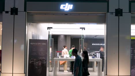 Los-Compradores-Exploran-Los-Productos-De-Drones-De-DJI-En-La-Tienda-Oficial-De-La-Compañía-En-Hong-Kong,-Mientras-Los-Peatones-Pasan-Por-La-Noche.