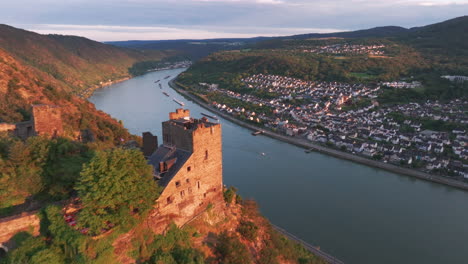 Fliegen-Sie-Vorbei-An-Einer-Burg-Am-Hang,-Die-Im-Orangefarbenen-Licht-Des-Sonnenuntergangs-Erstrahlt,-Und-Dem-Rhein-Und-Dem-Kleinen-Dorf-Im-Hintergrund