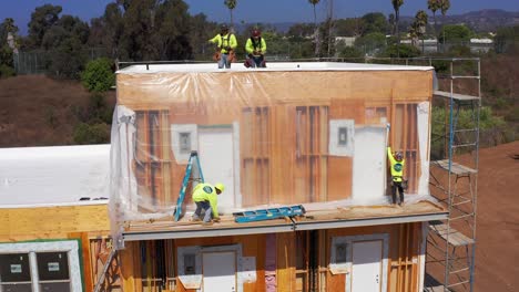 Slow-aerial-panning-shot-of-construction-crew-working-on-the-siding-of-a-housing-module-at-a-pre-fabricated-building-site-in-West-Los-Angeles,-California