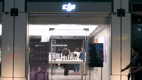 Customers-shop-at-the-Chinese-technology-company-DJI-official-store,-known-for-its-drone-product-line,-while-pedestrians-walk-past-the-frame-during-nighttime-in-Hong-Kong