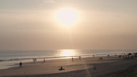 Sun-Setting-Over-Sandy-Seminyak-Beach-with-Tourists-Walking-by-the-Sea-Summertime-in-Bali,-Indonesia