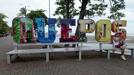 Young-man-sits-and-uses-his-phone-in-front-of-the-artistic-and-touristic-Quepos-location-sign-on-the-promenade-in-Costa-Rica