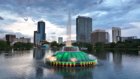 Lake-Eola-Park-in-downtown-Orlando,-featuring-the-iconic-fountain-illuminated-in-green,-with-the-city-skyline-and-a-rainbow-amphitheater-in-the-background