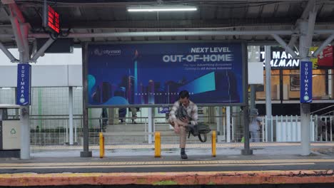 Un-Hombre-Esperando-En-La-Estación-De-Tren-De-Sudimara-Mirando-Su-Teléfono-Mientras-Estaba-Sentado-Bajo-Un-Gran-Cartel-Publicitario-En-Un-Día-Nublado