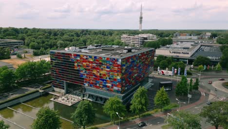 Drone-exterior-overview-of-the-colorful-Beeld-en-Geluid-building-at-Hilversum-Media-Park,-highlighting-its-unique-architecture-and-vibrant-glass-facade