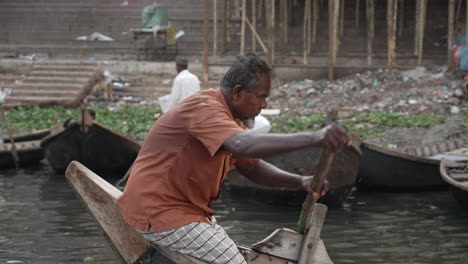 Mittlere-Aufnahme-Eines-Wassertaxi-Bootsführers,-Der-Ein-Traditionelles-Boot-Namens-Nouka-Entlang-Des-Buriganga-Flussufers-In-Dhaka-Paddelt,-Wobei-Im-Hintergrund-Andere-Noukas-Zu-Sehen-Sind