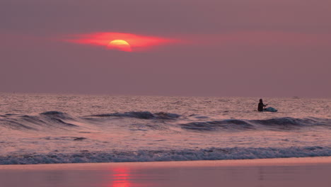 Burning-Sun-Setting-Over-Sea-in-Purple-Sky-Sunset,-Wave-Breaking-Over-Wet-Sand-Beach,-Silhouetted-Balinese-Man-Paddleboarding-on-Board-in-Background