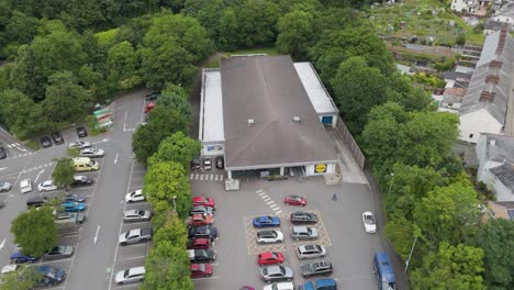 Drone-exterior-view-of-Lidl-store-and-parking-lot-in-Okehampton,-Devon-UK,-July-2024,-surrounded-by-greenery