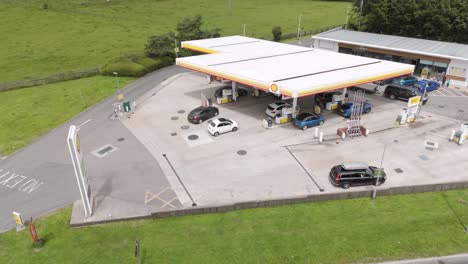 Aerial-view-of-cars-pulling-into-a-Shell-gas-petrol-station-in-Devon-UK,-surrounded-by-green-fields,-July-2024