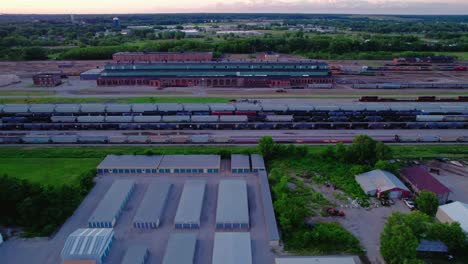 Aerial-view-of-a-rail-yard-with-trains,-storage-units,-greenery,-and-distant-horizon-at-sunset