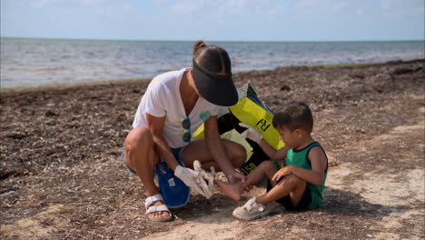 Zeitlupe-Eines-Jungen-Mexikanischen-Lateinamerikanischen-Jungen,-Dem-Seine-Mutter-Beim-Anziehen-Seiner-Turnschuhe-Hilft,-Während-Er-Am-Strand-Sitzt-Und-Sich-Freiwillig-Bei-Der-Strandreinigung-In-Cancun-Engagiert.