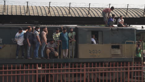 Los-Niños-Se-Sientan-Peligrosamente-En-El-Techo-De-Un-Tren-Que-Pasa-Por-La-Estación-De-Tren-Del-Aeropuerto-En-Dhaka,-Bangladesh