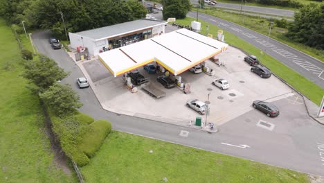 Drone-view-of-cars-at-a-Shell-petrol-gas-station-in-Devon-UK,-surrounded-by-greenery-and-roads,-July-2024