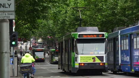 Straßenbahnen-Fahren-Entlang-Der-Von-Bäumen-Gesäumten-Swanston-Street,-Während-Essenslieferanten-Auf-Dem-Fahrradweg-Neben-Den-Straßenbahnschienen-In-Melbournes-Geschäftigem-Stadtzentrum-Fahren