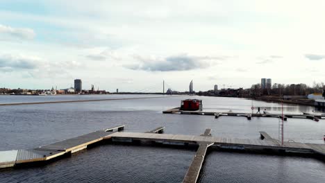 Ein-Langer,-Hölzerner-Steg-Ragt-In-Ein-Großes,-Ruhiges-Gewässer-Hinein,-Der-Fluss-Daugava-An-Einem-Sonnigen-Tag,-Ruhiges-Gewässer-Mit-Ein-Paar-Booten,-Die-In-Der-Marina-Angedockt-Sind