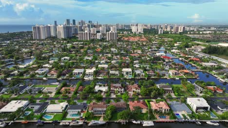 Aerial-view-of-a-coastal-suburban-neighborhood-with-white-roofed-houses,-lush-greenery,-and-canals,-transitioning-to-high-rise-buildings-in-the-background