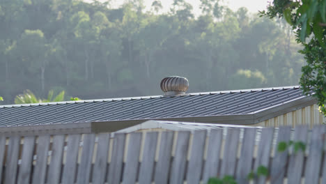 Pan-Left,-A-metal-shed-with-a-whirlybird-on-its-roof-in-the-early-morning-sunlight-with-mountain-terrain-in-background,-Townsville,-Queensland,-Australia