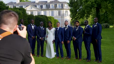 wedding-photo-shoot,-view-of-the-photographer-from-the-back-on-the-left-and-the-very-pretty-black-skinned-bride-with-her-husband-and-all-the-groomsmen,-on-a-lawn-with-the-chateau-in-the-background