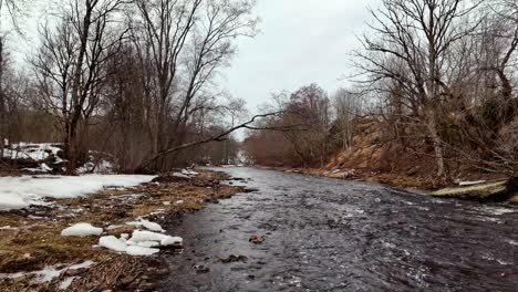 Ein-Fluss-Mit-Schneebedecktem-Ufer-Und-Bäumen-Im-Hintergrund