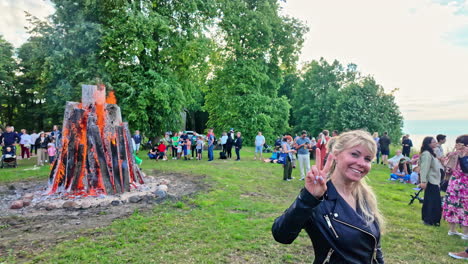 Süße-Blonde-Frau-Hält-Lächelnd-Ein-Peace-And-Rock-Schild-Hoch,-Während-Hinter-Ihr-Während-Des-Festivals-Das-Feuer-Wütet